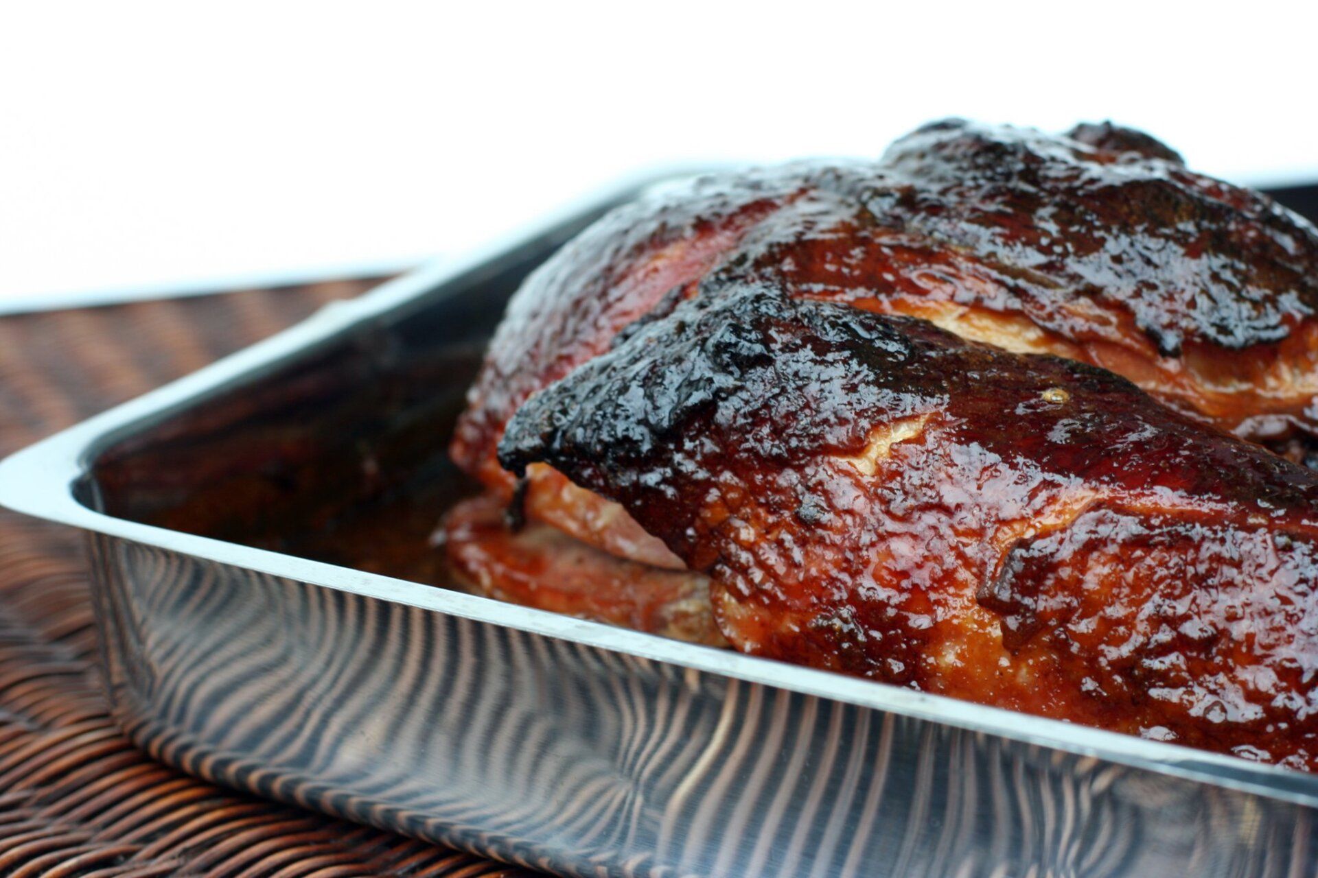 A glazed, roasted piece of meat with a dark, caramelized crust sitting in a metallic baking pan on a textured surface.