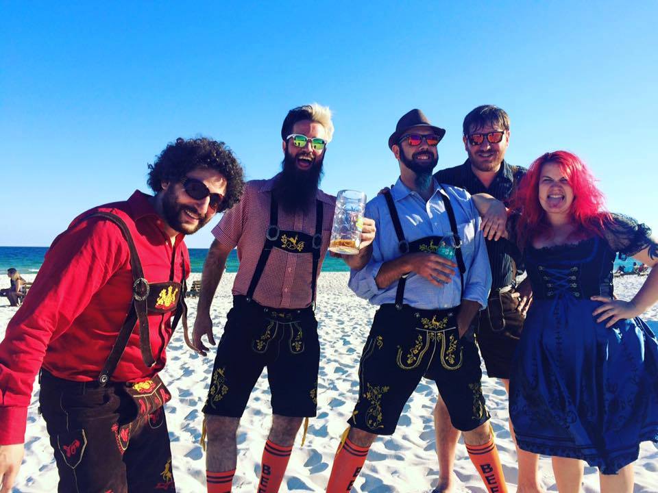 Four people in traditional Bavarian clothing pose on a sunny white sand beach.