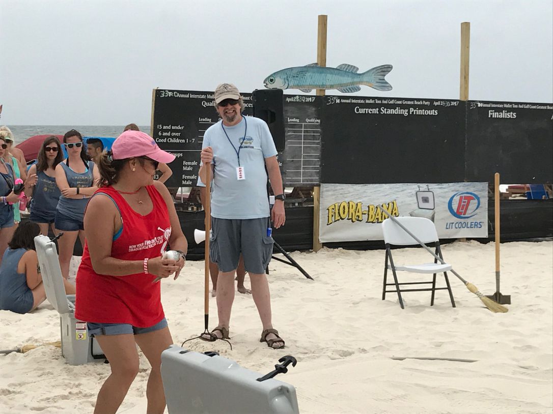 A person in a red tank top stands on a sandy beach during an event, facing a person holding a staff near a score board.