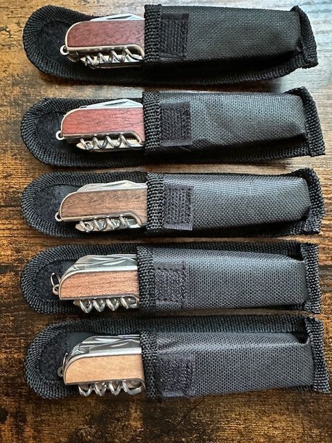 Five pocket knives in black fabric sheaths, each with wooden handles and corkscrews, on a wooden surface.