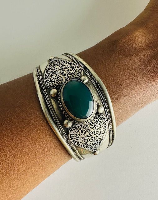 Silver cuff bracelet with green oval stone, worn on a wrist. Intricate filigree design.