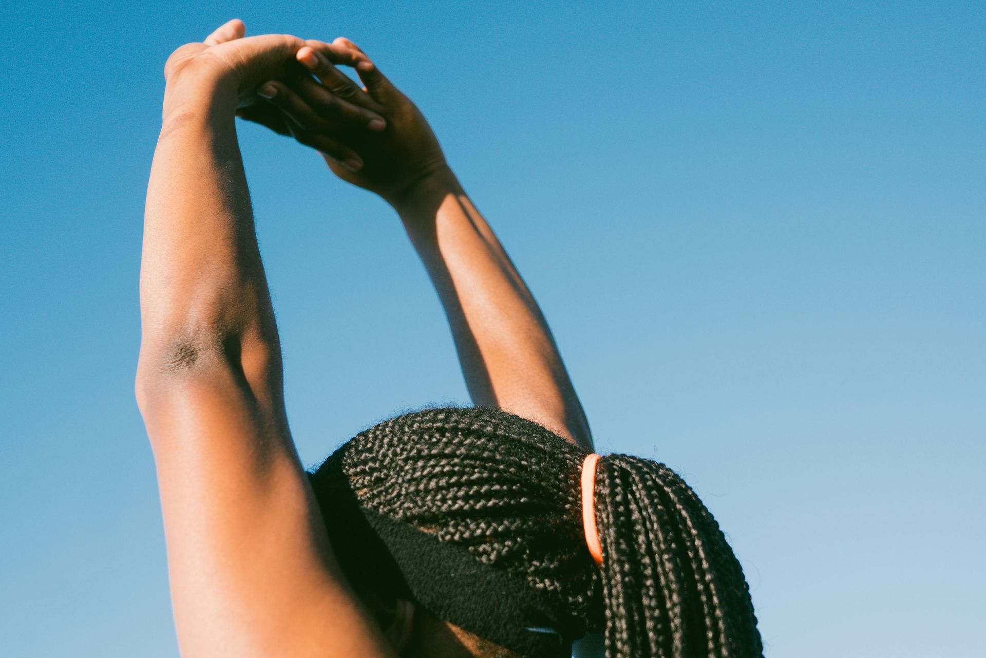 Person with braided hair stretching arms overhead against a blue sky.