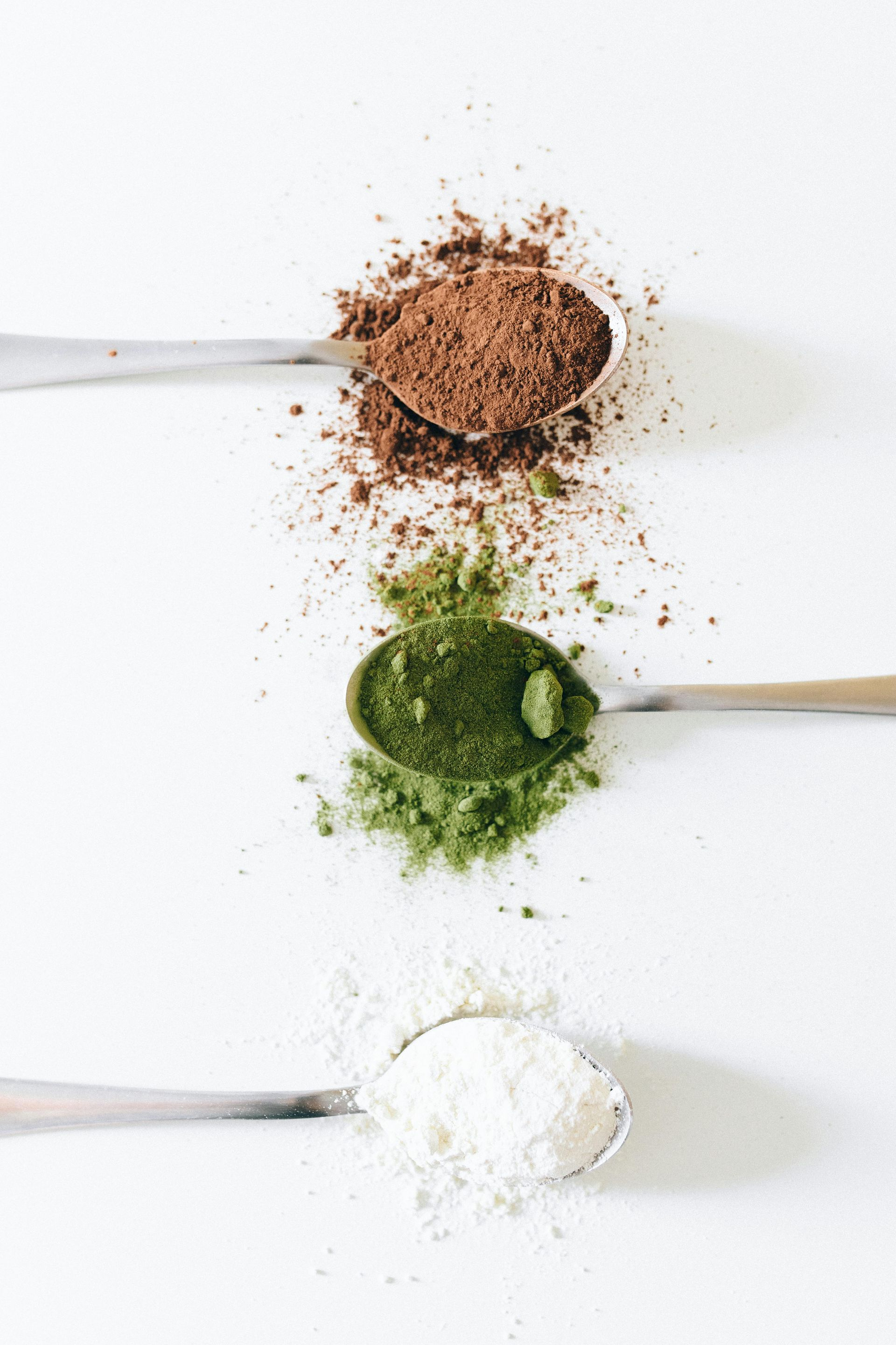 Spoons with cocoa, green powder, and white powder, aligned vertically on a white surface. Text: