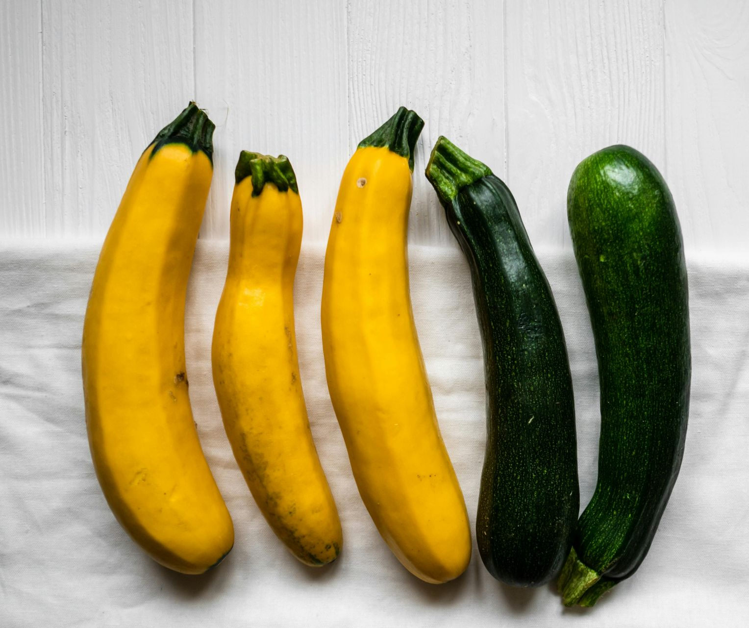 Five yellow and green zucchini arranged on a white, textured surface.