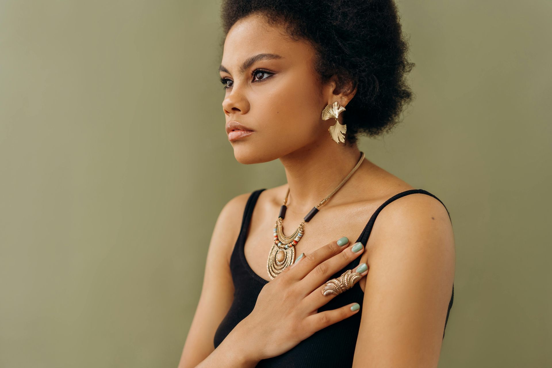 Woman wearing gold jewelry, hand on chest, looking away from the camera. Black tank top, green background.