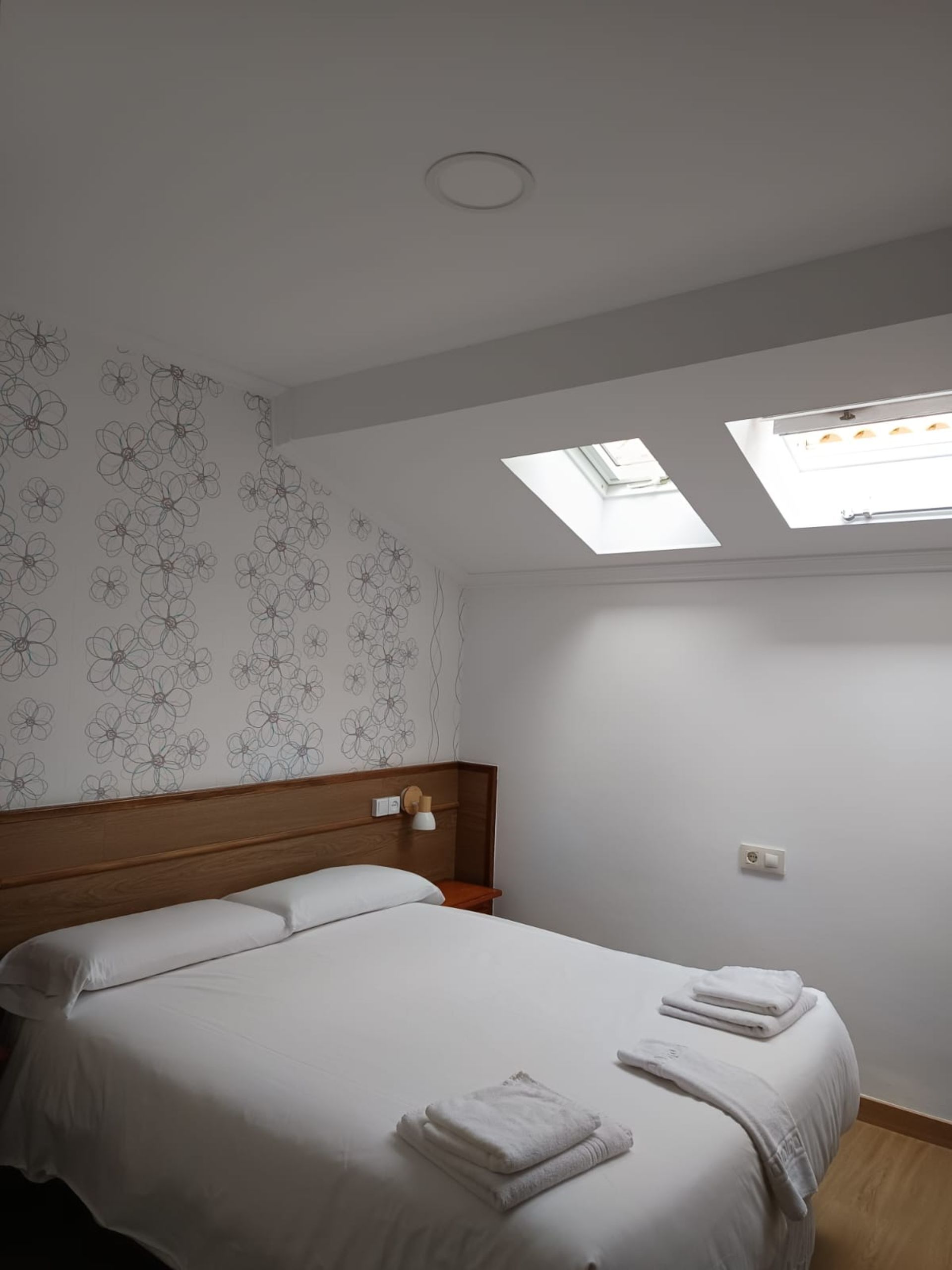Habitación pequeña con cama doble, papel pintado estampado y ventanas de techo.