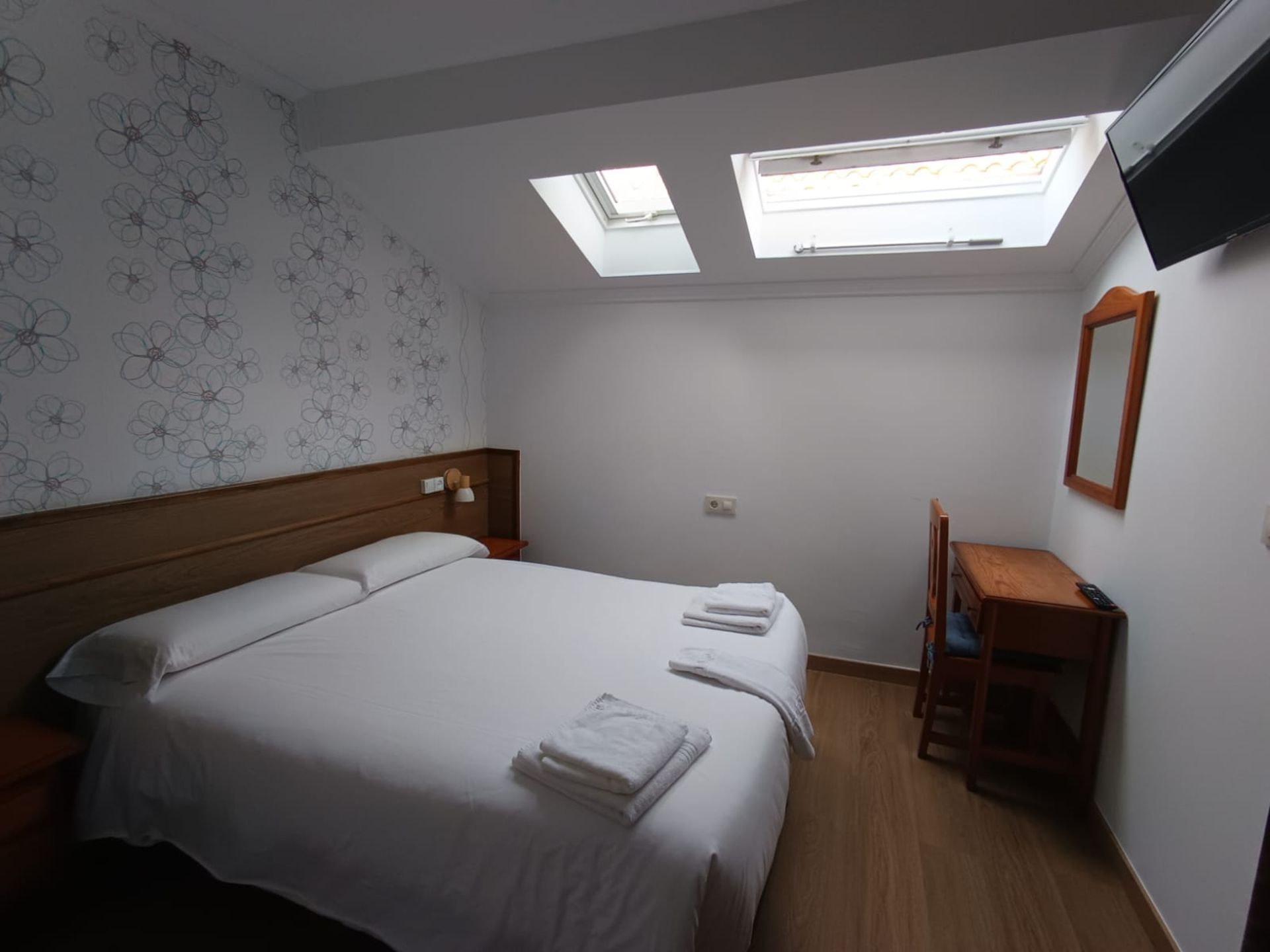 Habitación pequeña con cama doble, escritorio y silla de madera, paredes blancas y ventanas de techo.