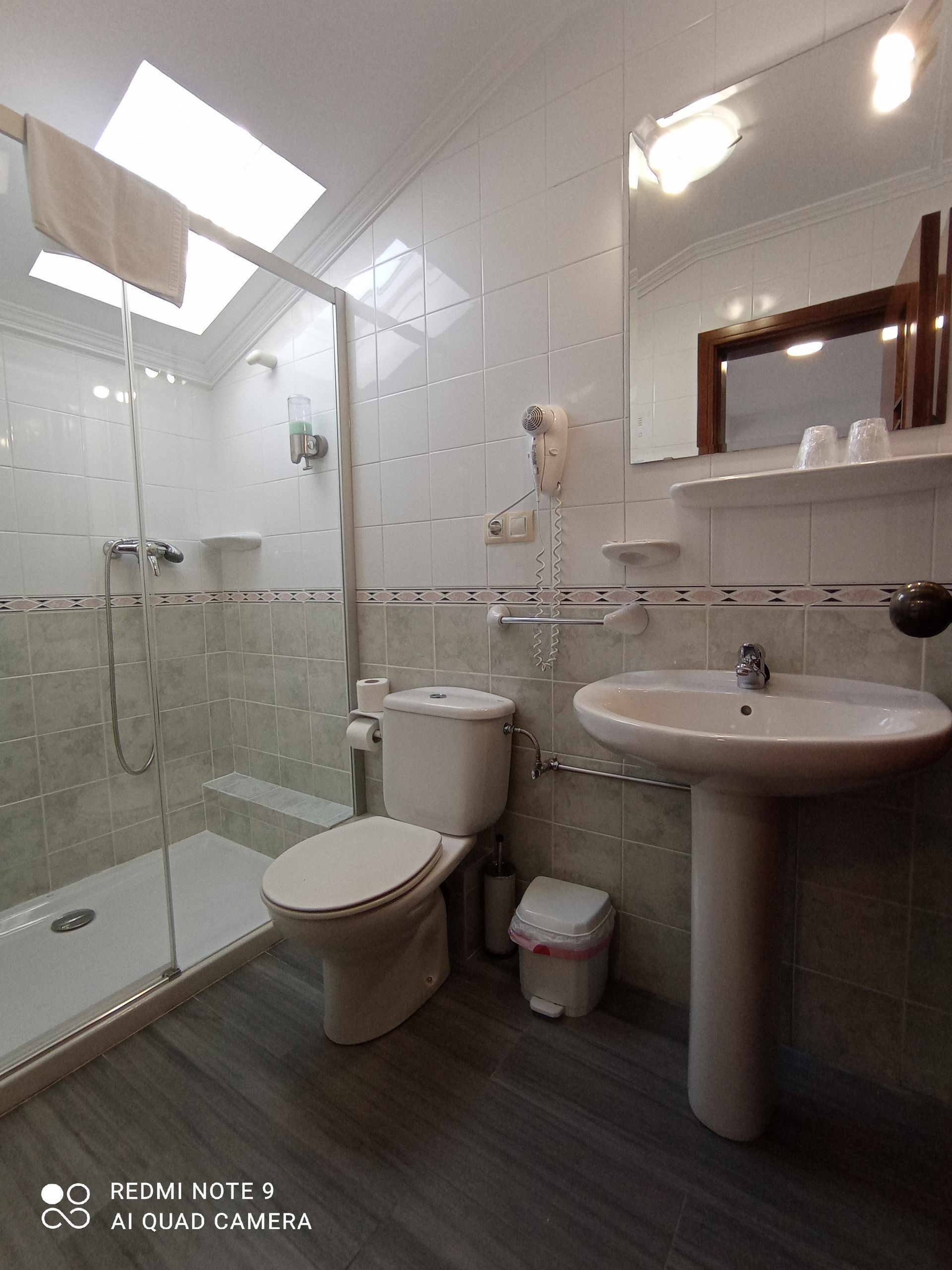 Baño pequeño con azulejos blancos, ducha, inodoro, bidé, lavabo, espejo e iluminación a través de una claraboya.