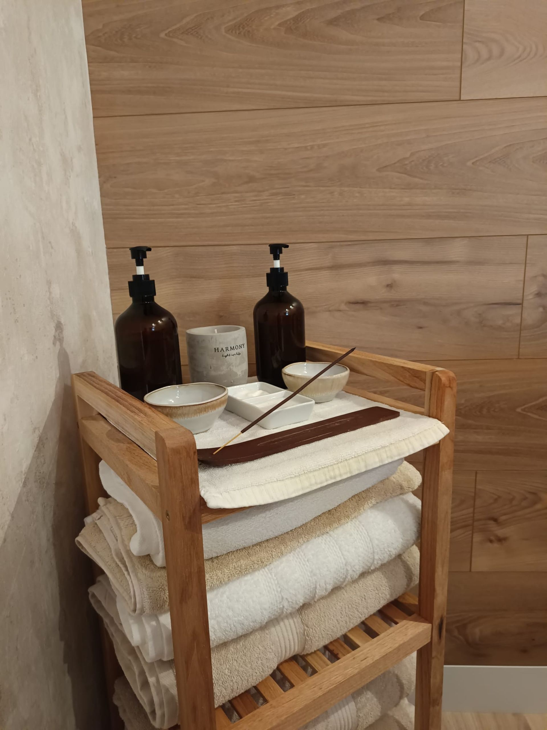 Estantería de madera para spa con toallas dobladas, botellas y una pequeña bandeja en un rincón alicatado del baño.