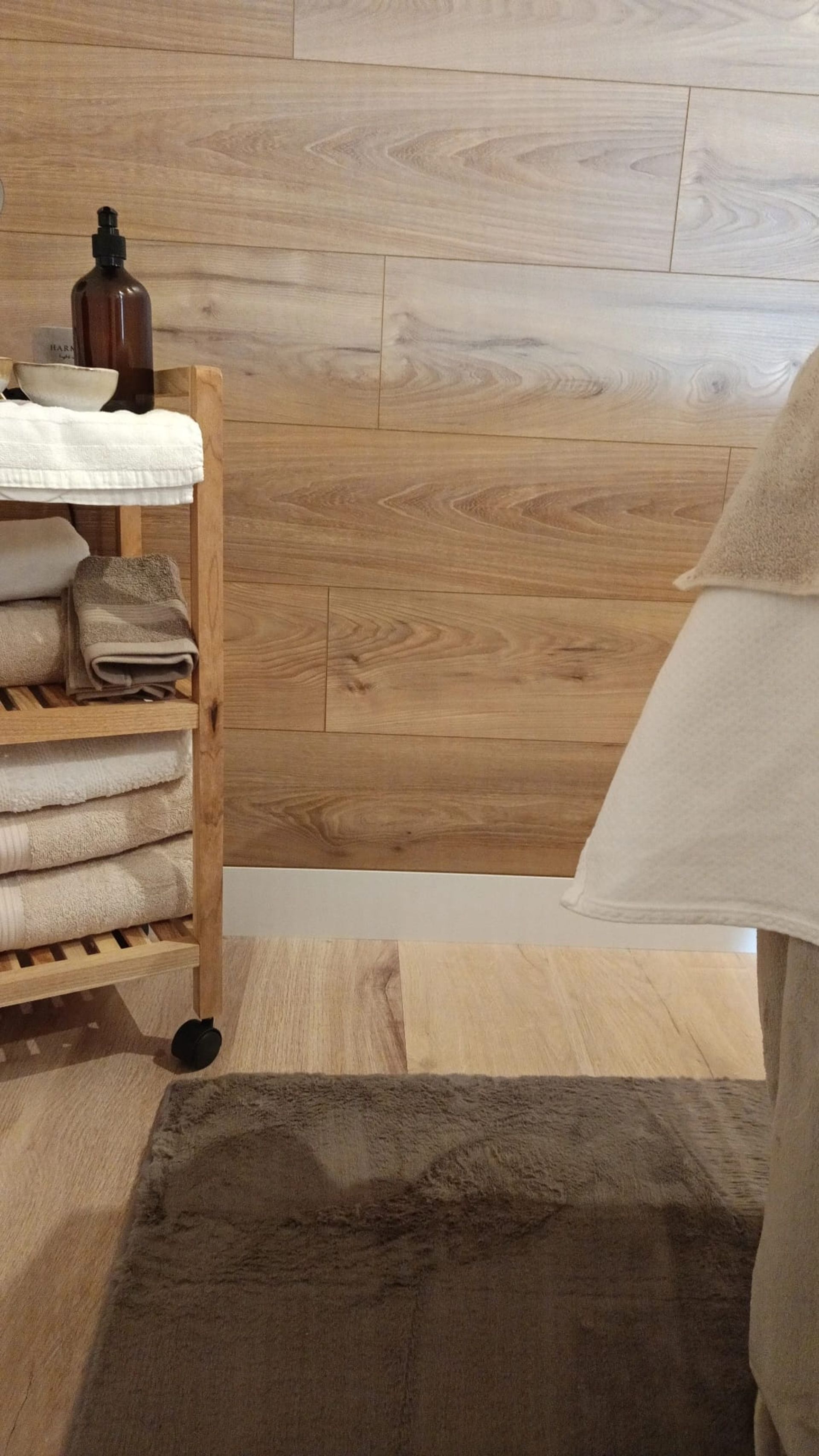 Baño de madera con carrito de mimbre, toallas beige, alfombrilla de baño y borde blanco para bañera o cama.