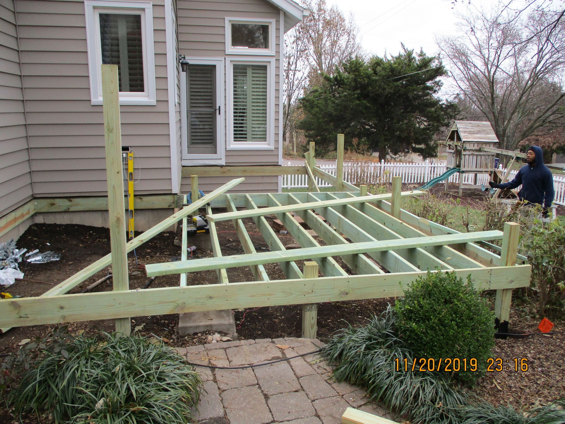 Raised wooden deck frame under construction beside a house, with a worker using a tool on the right.