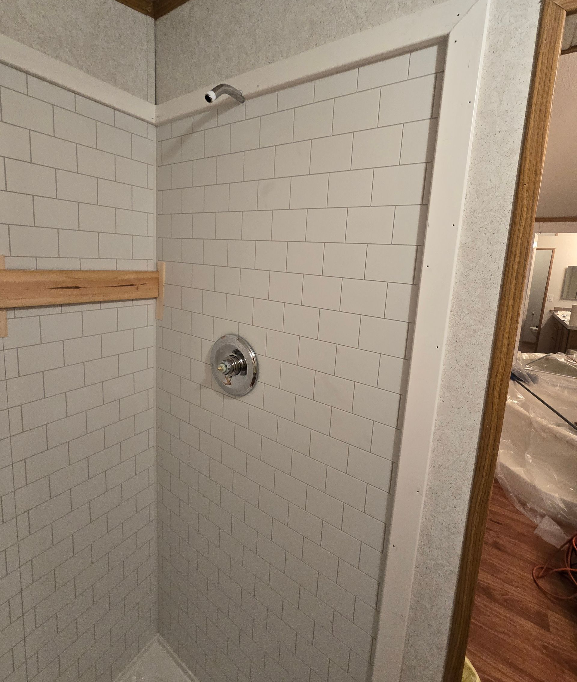 White-tiled shower stall with chrome showerhead and built-in corner shelf; bathroom doorway visible.
