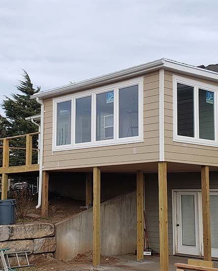 Elevated beige house on wooden stilts with large windows and a white door on a sloped lot