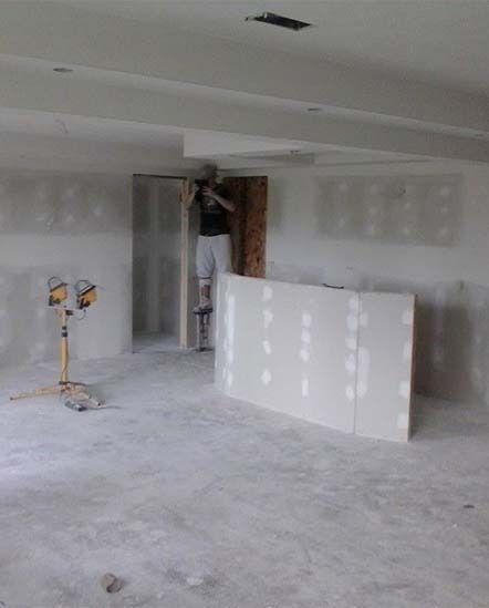 Unfinished drywall interior with a worker near a doorway and a large drywall panel in the foreground.