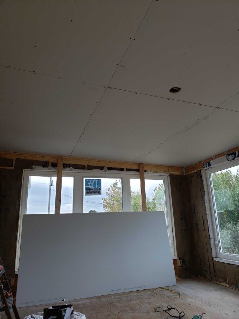 Partially finished room with white ceiling panels, exposed walls, large windows, and a white board leaning inside.