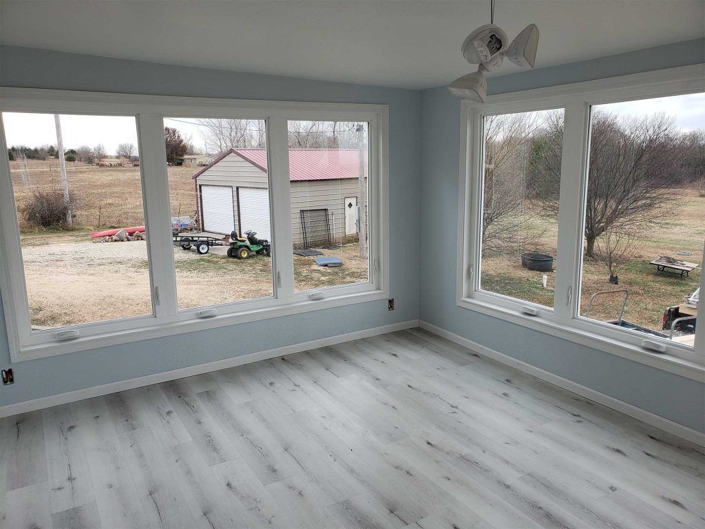 Empty sunroom with light gray floors, pale blue walls, and large windows overlooking a yard and shed.