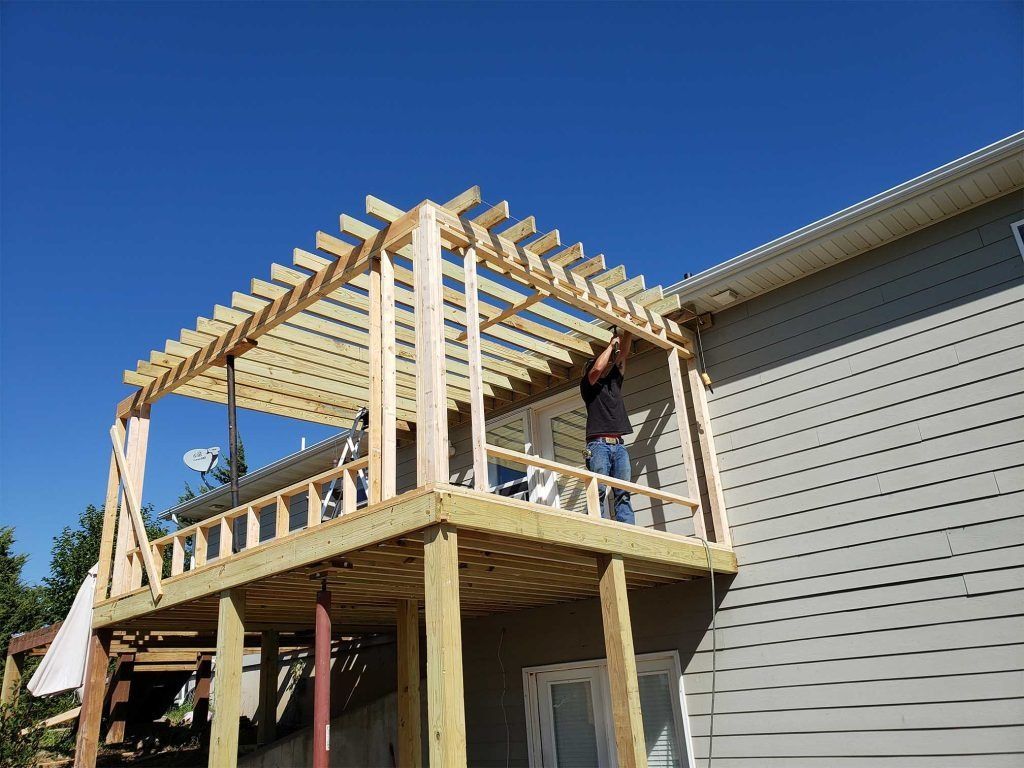 Partially built wooden deck and pergola attached to a gray house, with two workers on the structure.