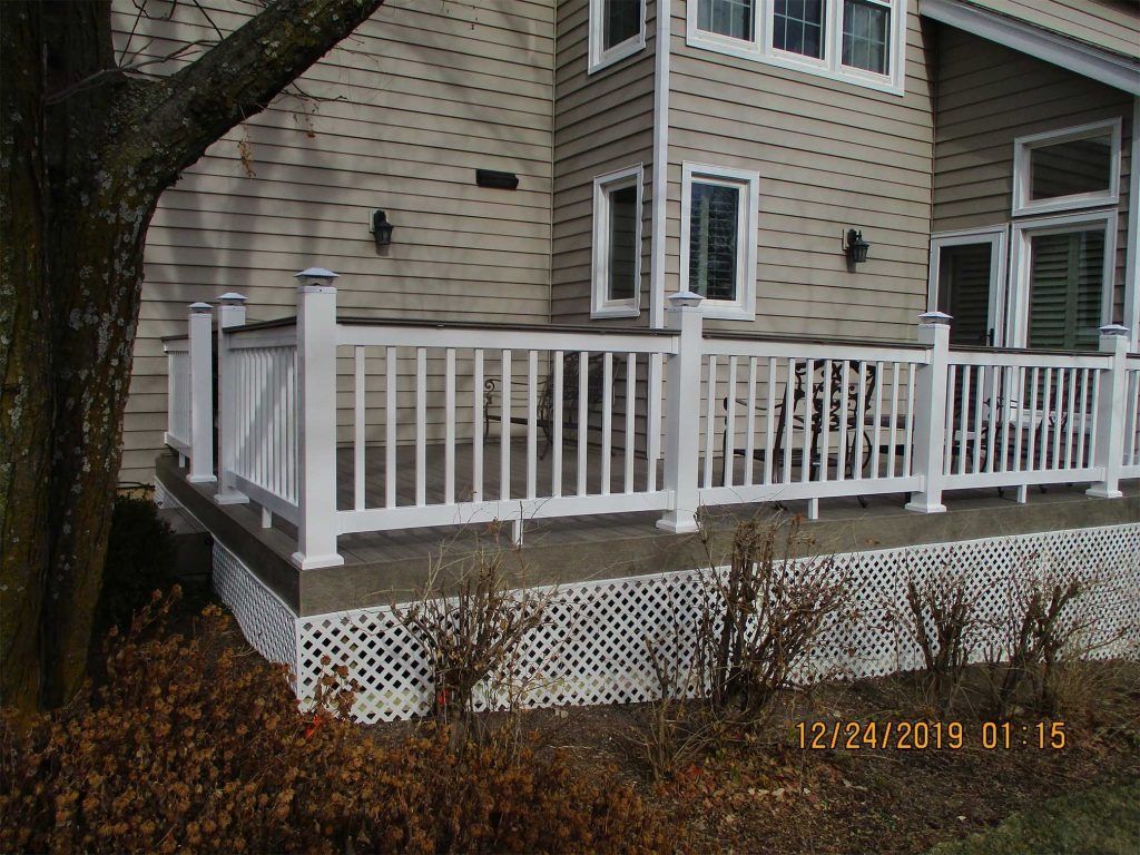 White wooden deck with railing attached to a beige house, beside a large tree and shrubs.