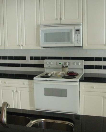 White kitchen with a stove, microwave, black tile backsplash, and sink in the foreground