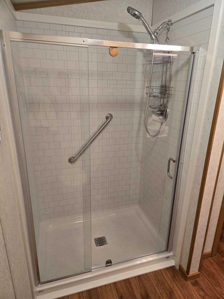 Accessible walk-in shower with sliding glass door, chrome grab bar, built-in caddy, and tiled walls.