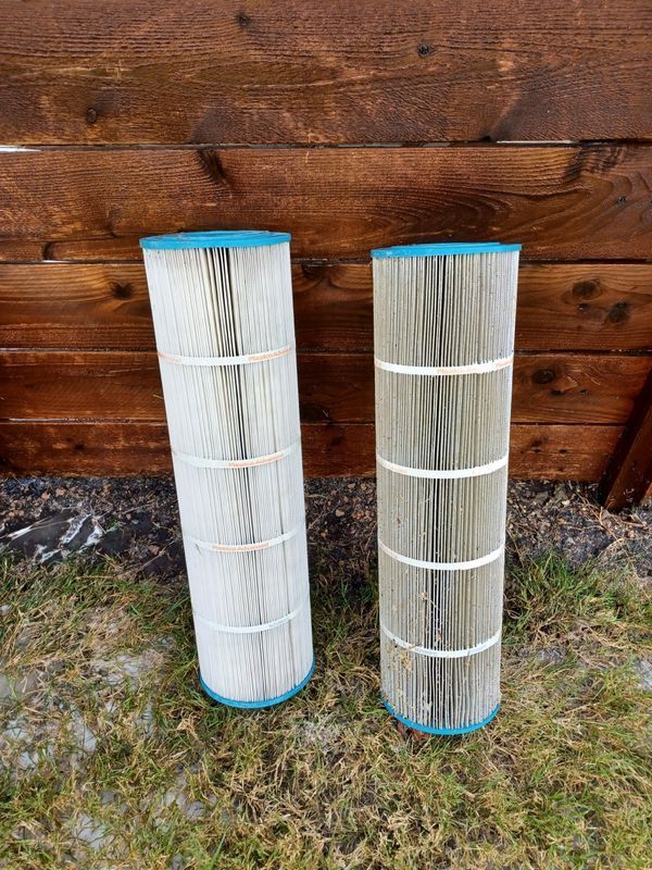 Two tall, cylindrical pool filter cartridges, one clean and white and one used and darkened, stand on grass by a fence.