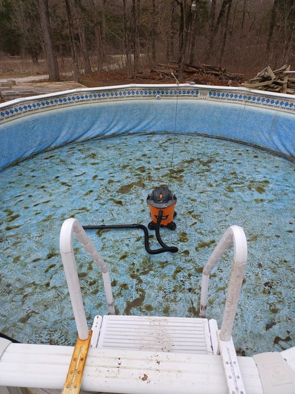 An orange wet-dry vacuum sits in the center of an empty, debris-covered above-ground swimming pool.