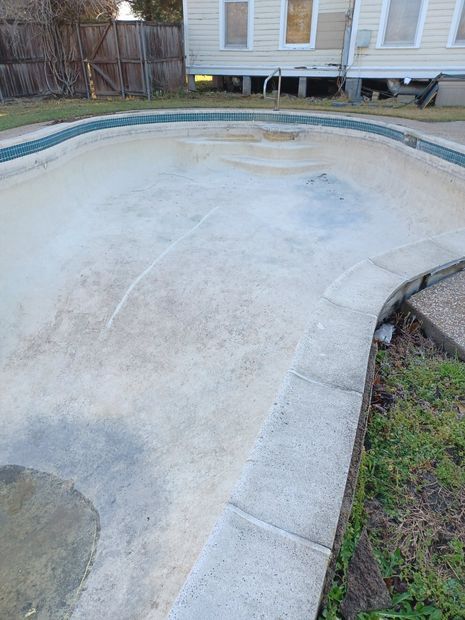 A drained, empty swimming pool with a light-colored plaster basin and blue tiled trim, situated next to a house.