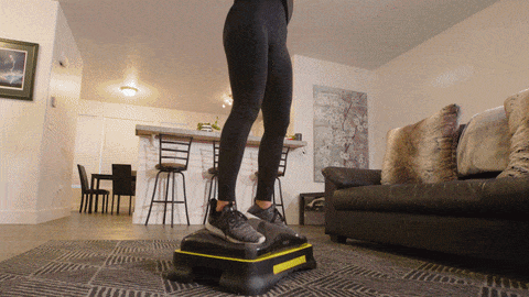 Person standing on a black-and-yellow exercise platform in a living room, with sofa and bar stools nearby