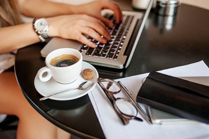 Hands typing on a laptop beside coffee, eyeglasses, and a notebook on a table