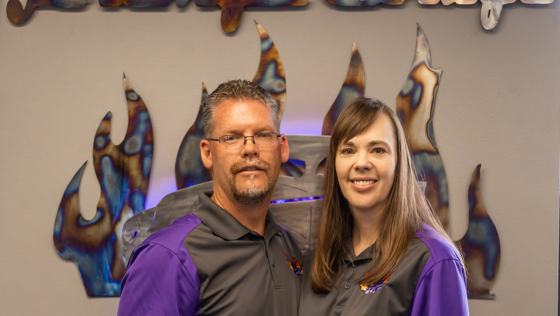 Couple in matching purple and gray shirts, standing in front of a flame-shaped metal art piece.