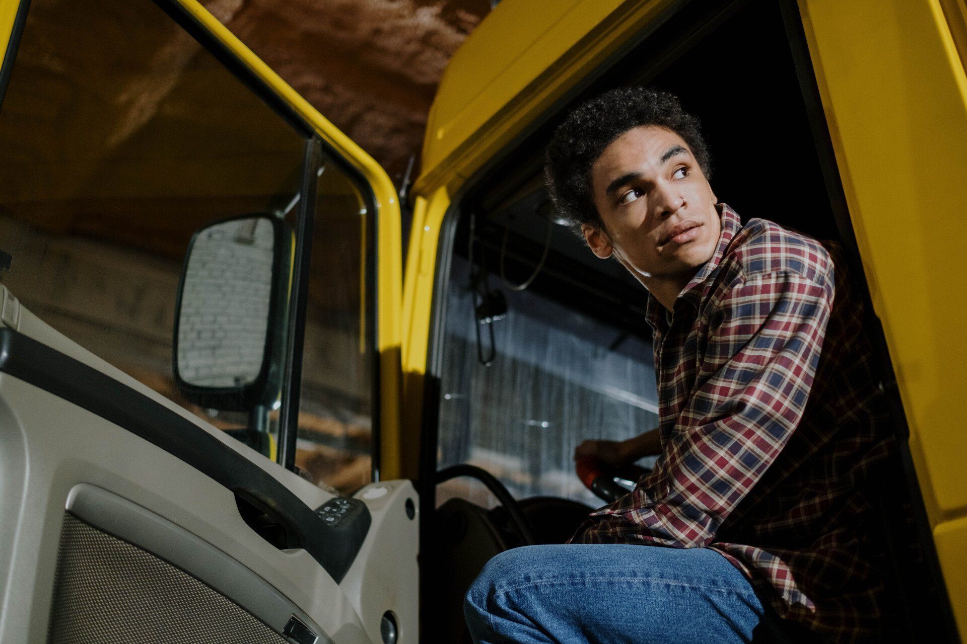 A person in a plaid shirt sits inside the cab of a yellow truck, looking over their shoulder toward the open door.
