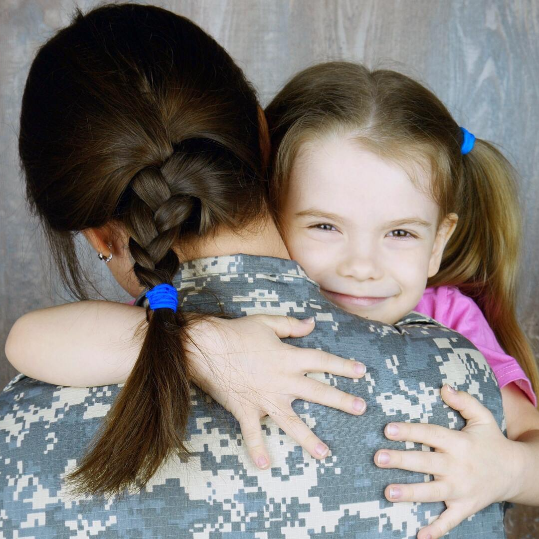 A young child in a pink shirt hugging a person wearing a digital camouflage military uniform.