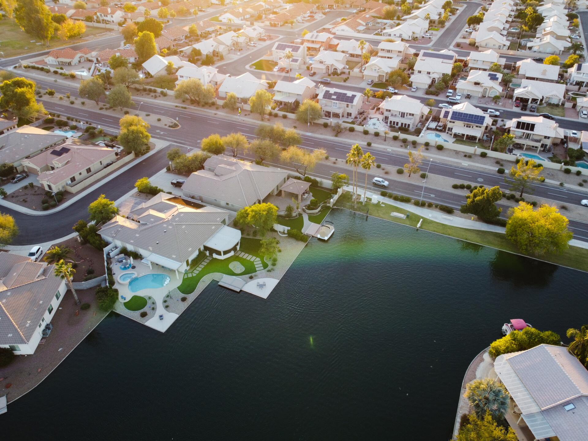 Aerial view of suburban homes with pools bordering a lake in a desert residential community at sunset.