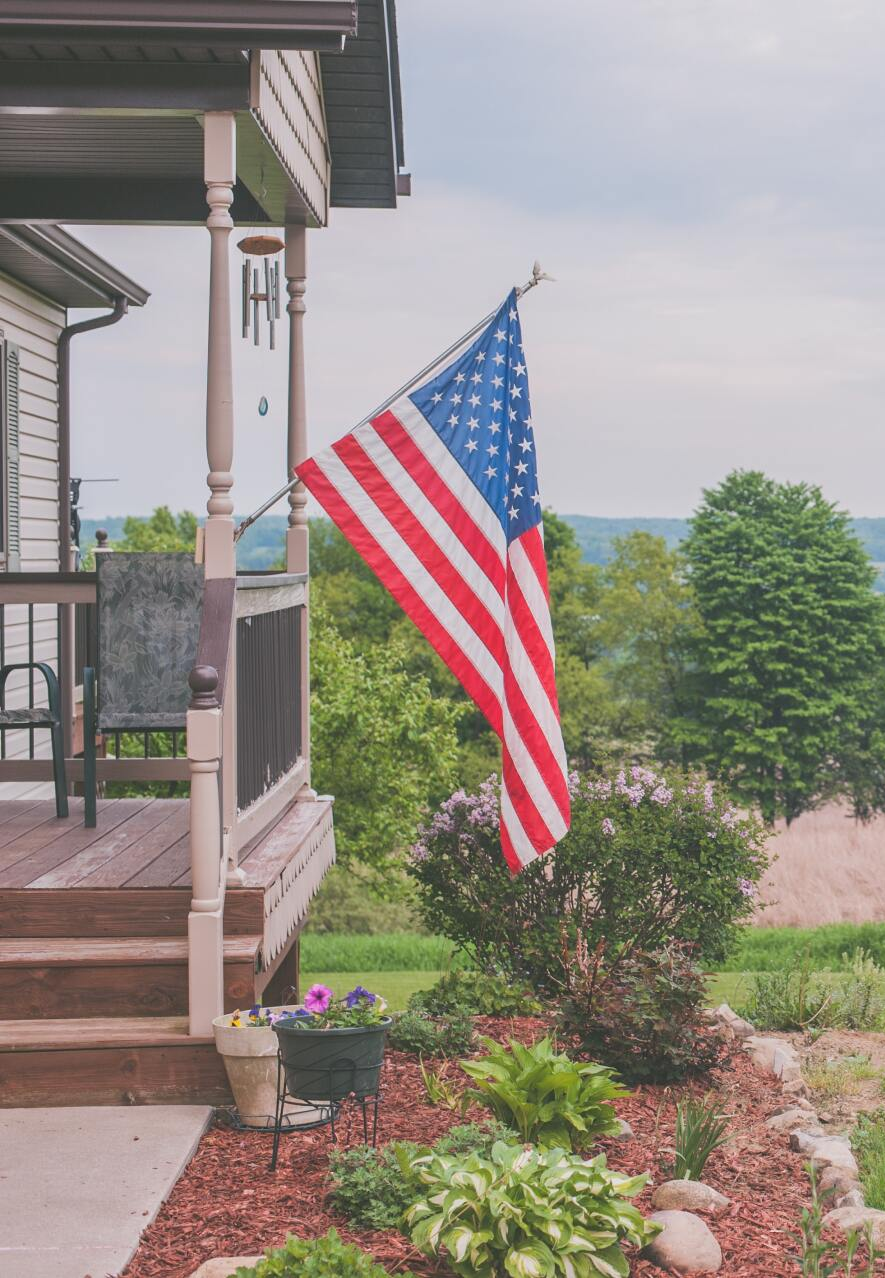 An American flag hangs from the porch of a house overlooking a green landscape with trees and a cloudy sky.