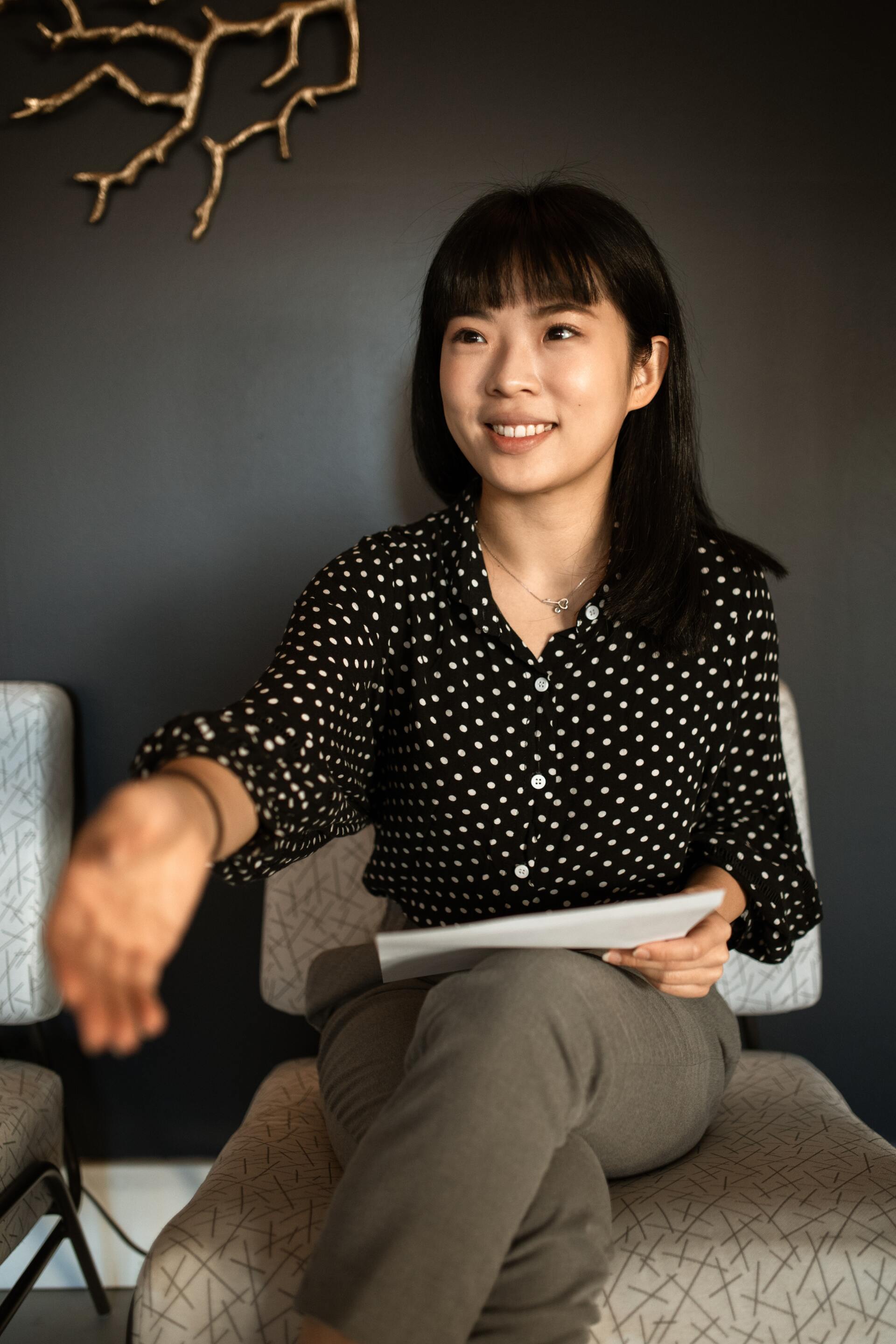 A smiling person in a polka-dot blouse sits in a chair, holding papers and reaching out with one hand against a gray wall.