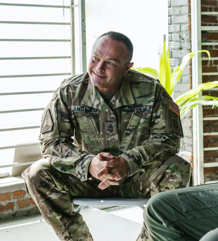 A person in a camouflage military uniform sits with hands clasped, smiling while looking toward someone off-camera.