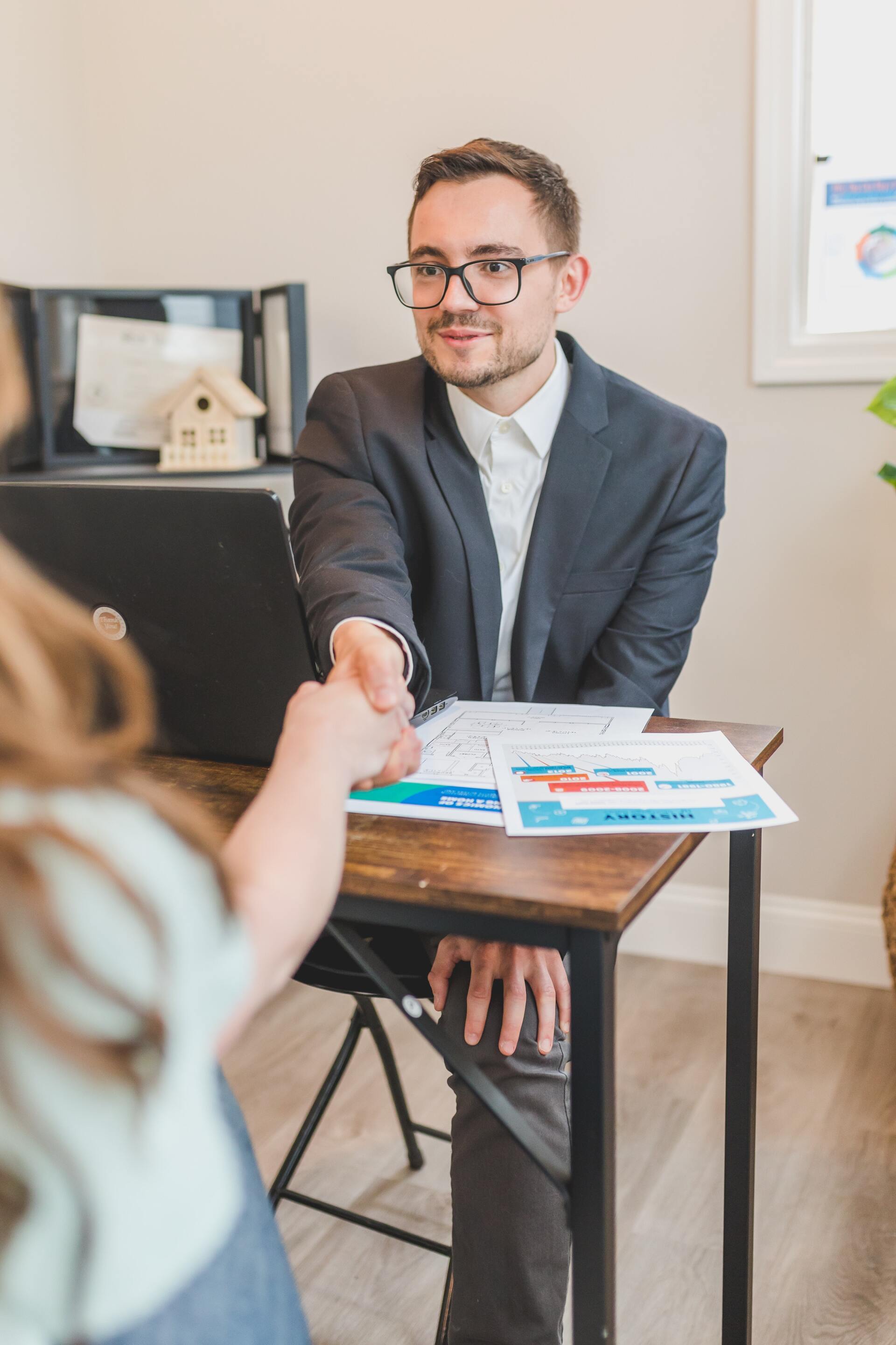 A professional in a suit shakes hands with a client across a desk in an office setting.