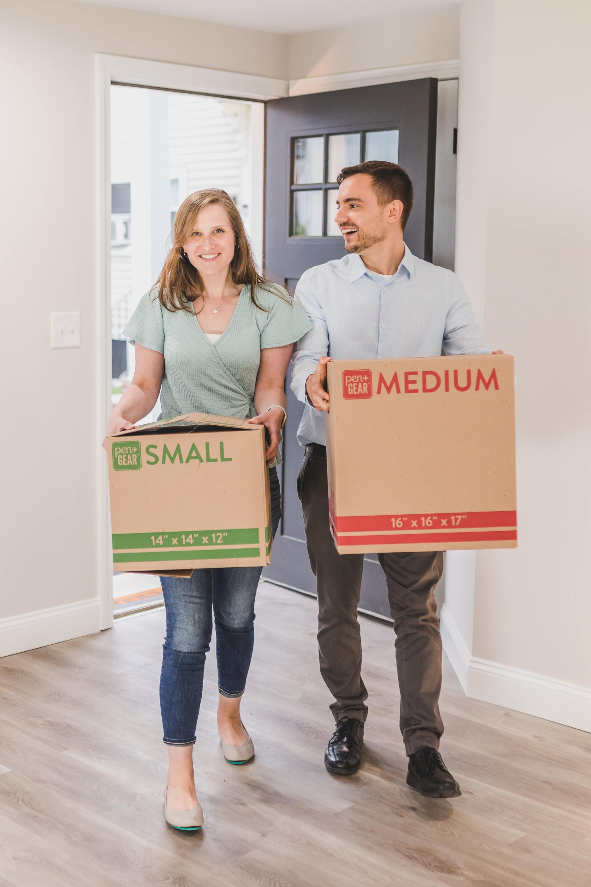 A couple smiling and walking into a new home while carrying a small and a medium-sized cardboard moving box.