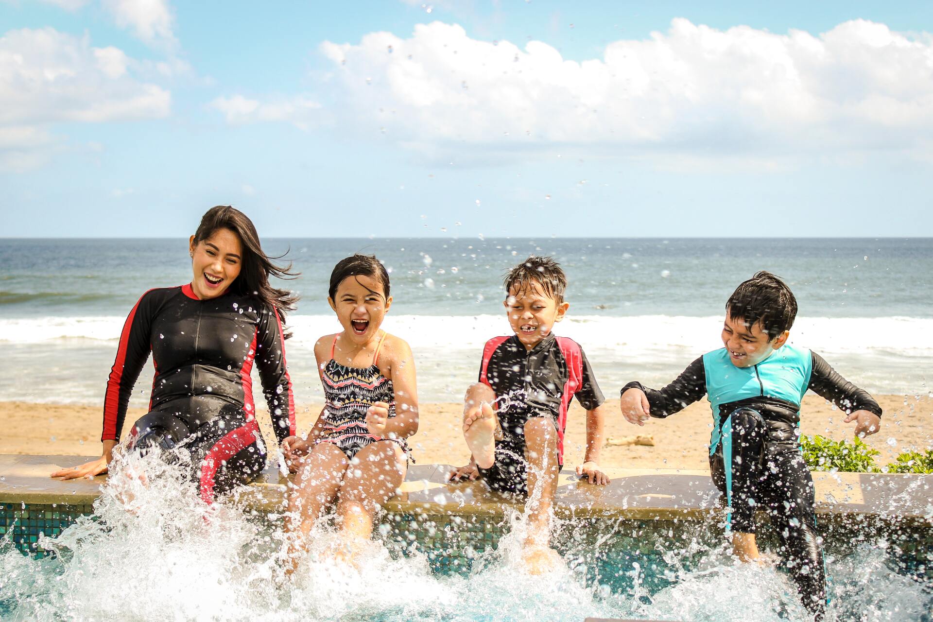 A person and three children wearing swimsuits splash water from a pool edge with the beach and ocean in the background.