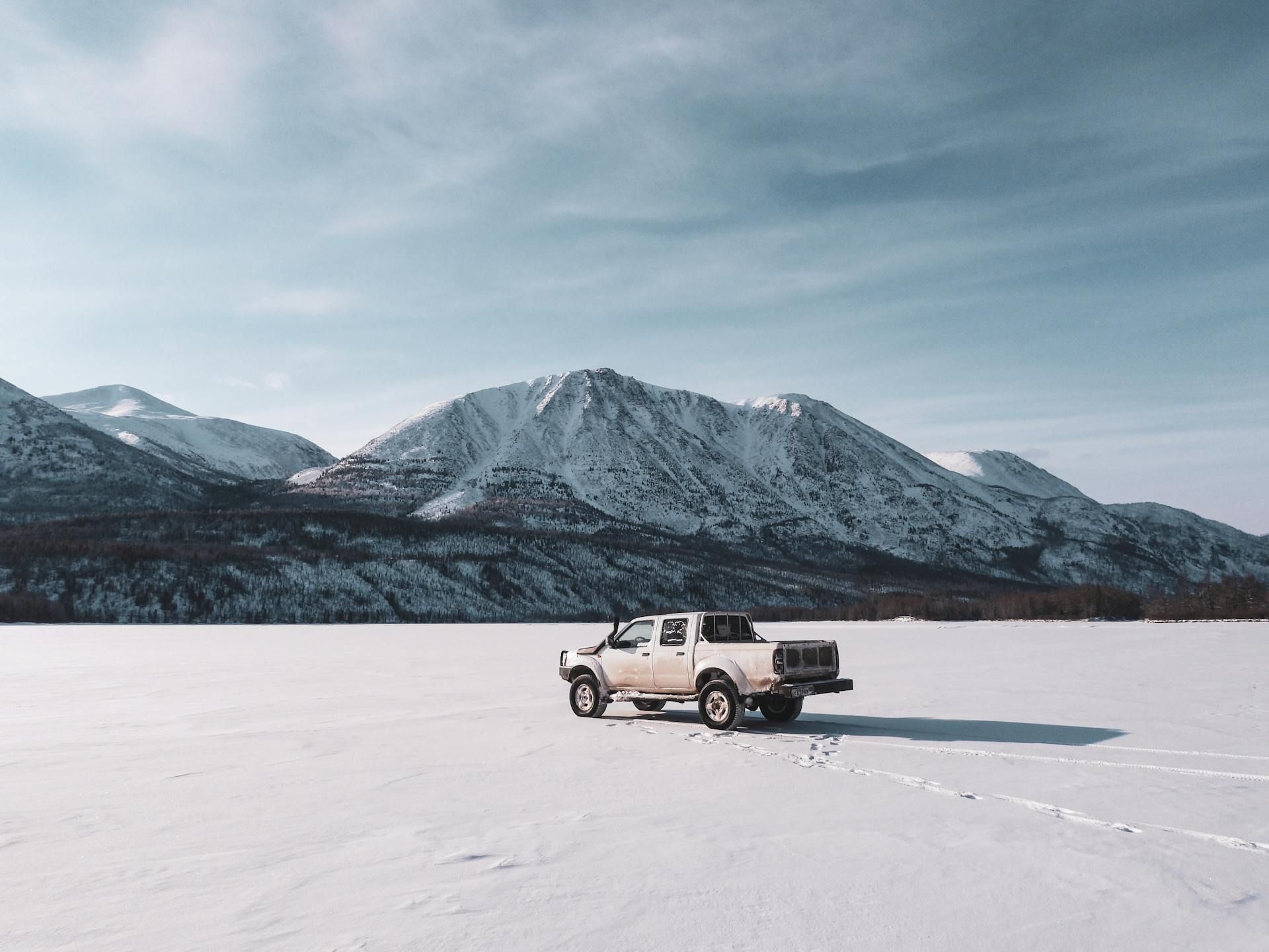 A white truck is parked in the middle of a snow covered field with mountains in the background.