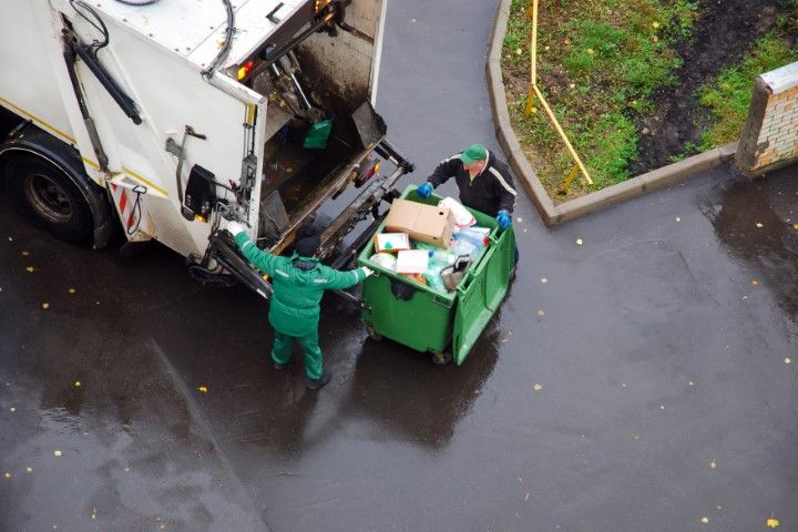 Garbage truck workers empty a green dumpster onto a wet street near a curb.