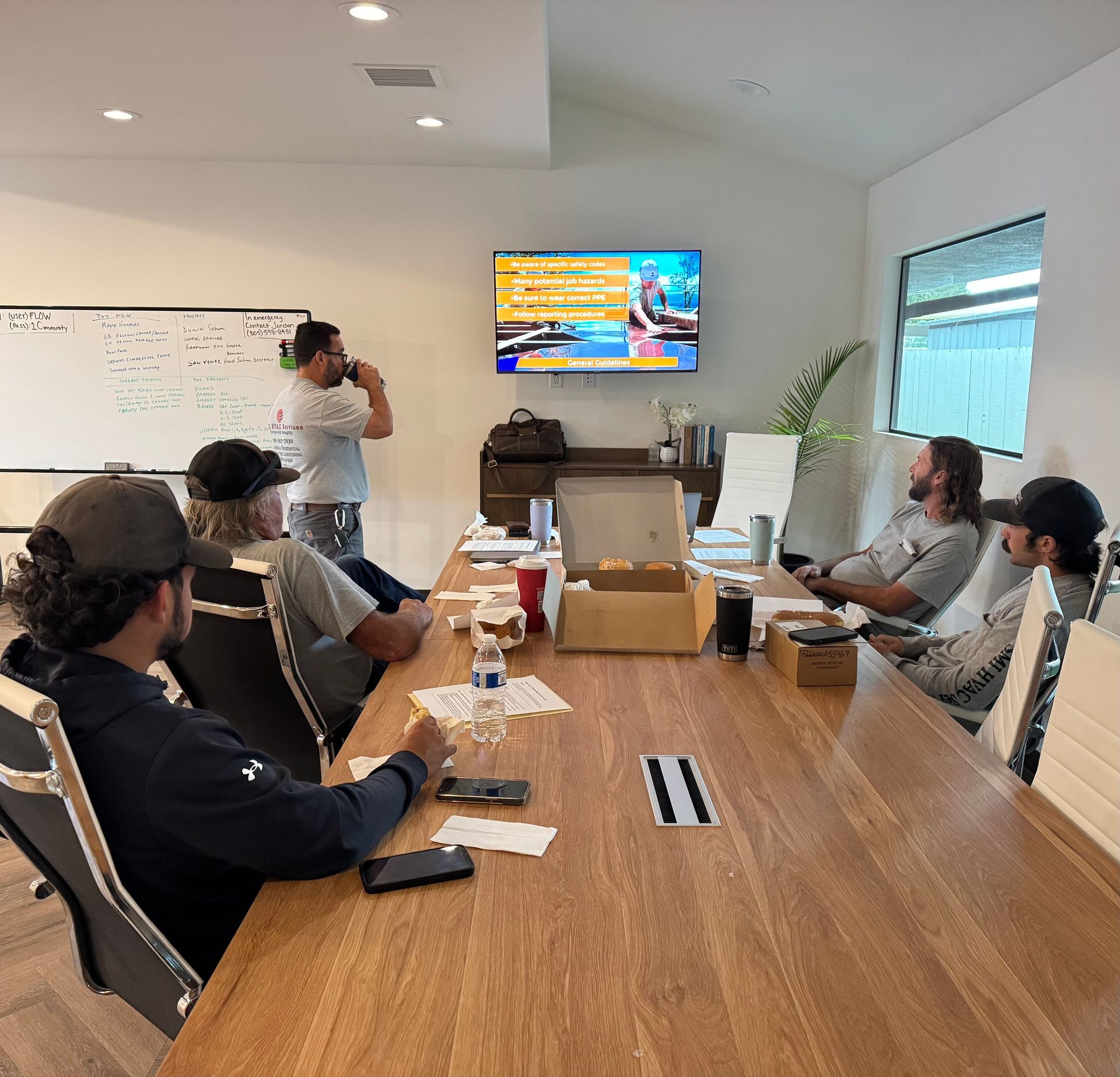 Group of people in a meeting room, looking at a TV screen while one man stands up holding a cup.