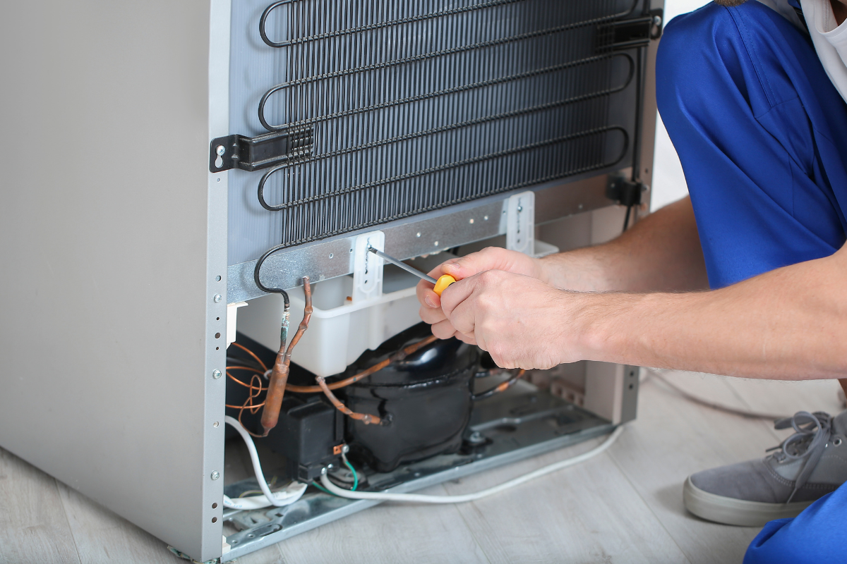 Person repairs refrigerator's back with screwdriver, crouching. Wires and compressor visible. Light-colored cabinet.