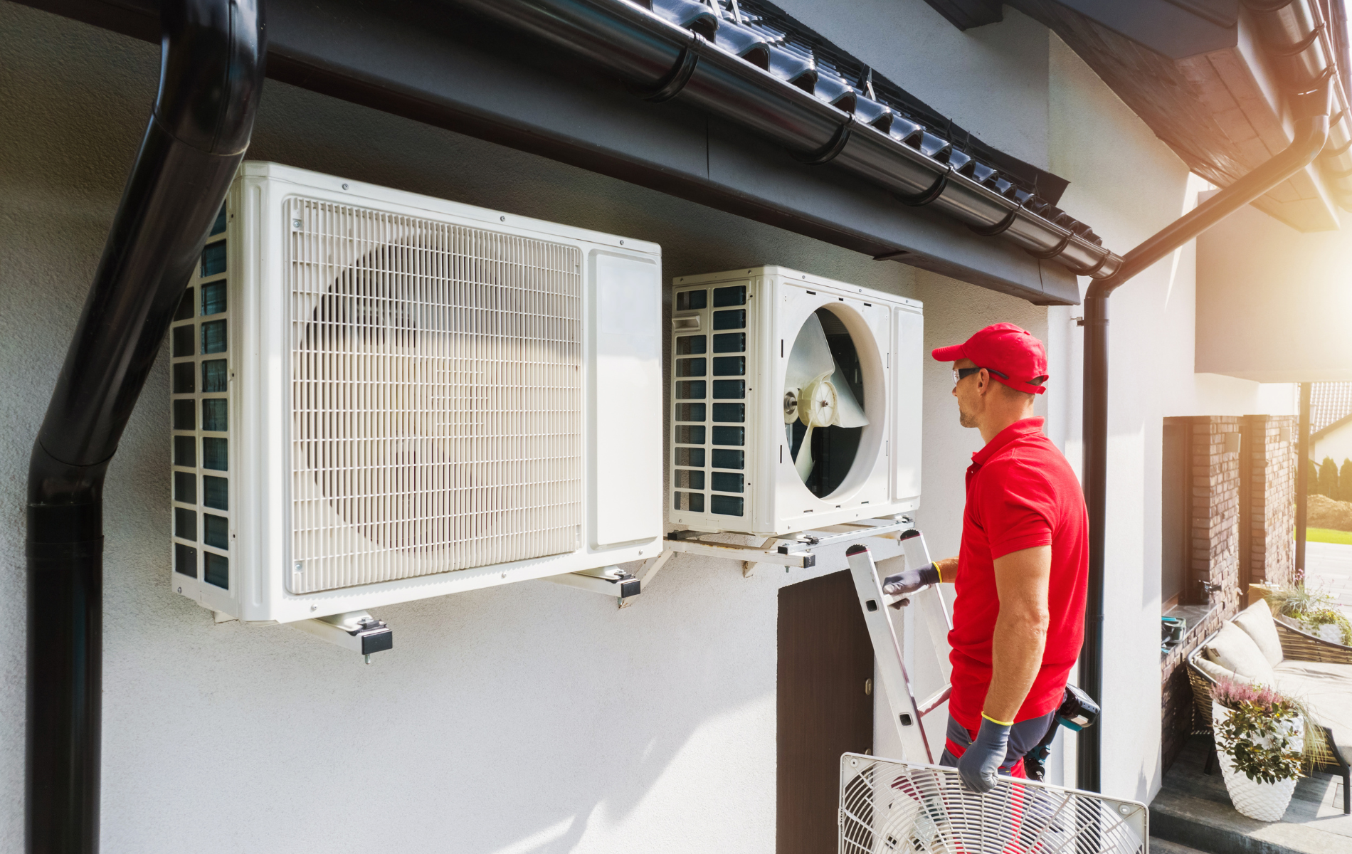 HVAC technician in red uniform installing or servicing two white air conditioning units mounted on a house.