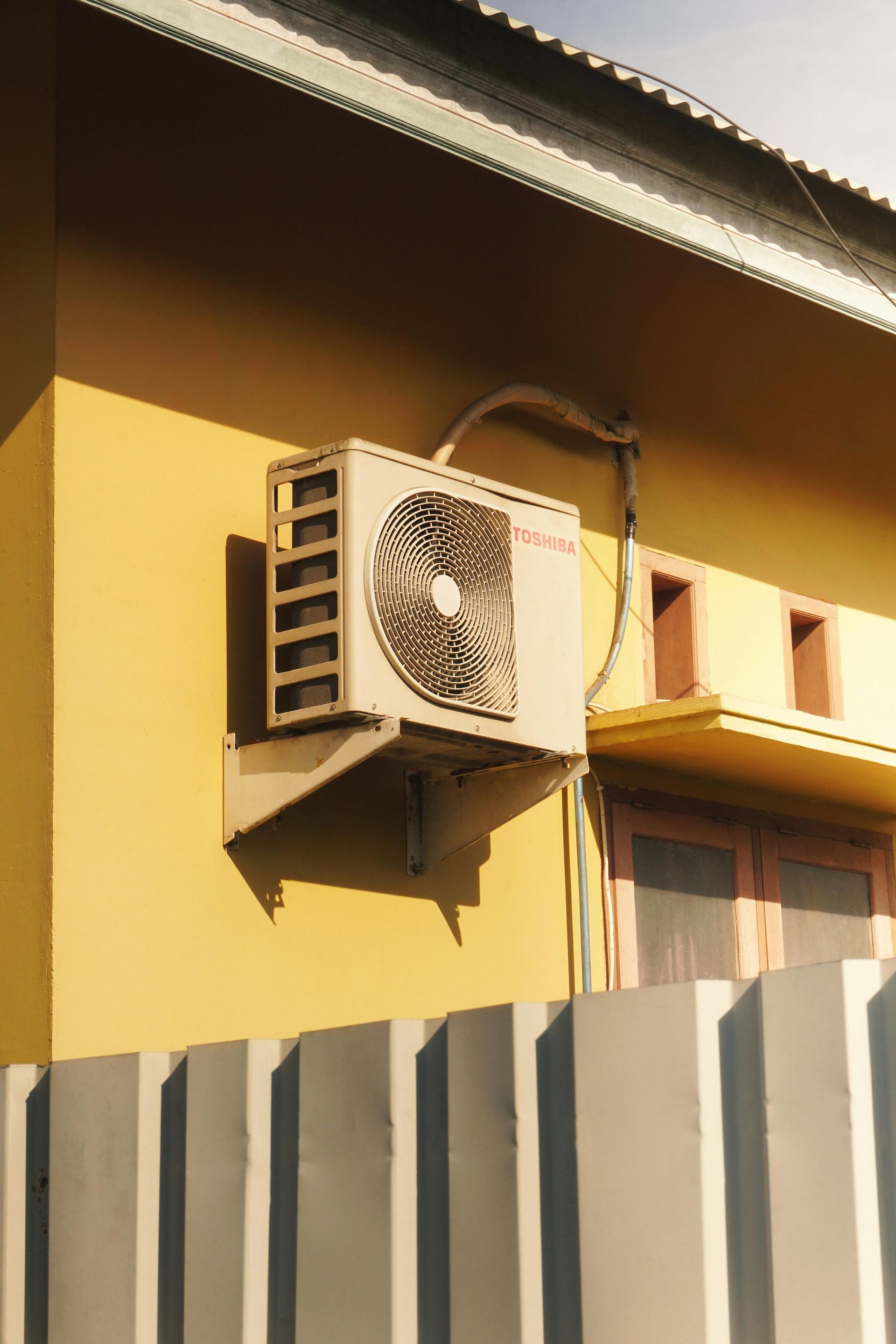 Air conditioning unit mounted on a yellow exterior wall above a gray fence.
