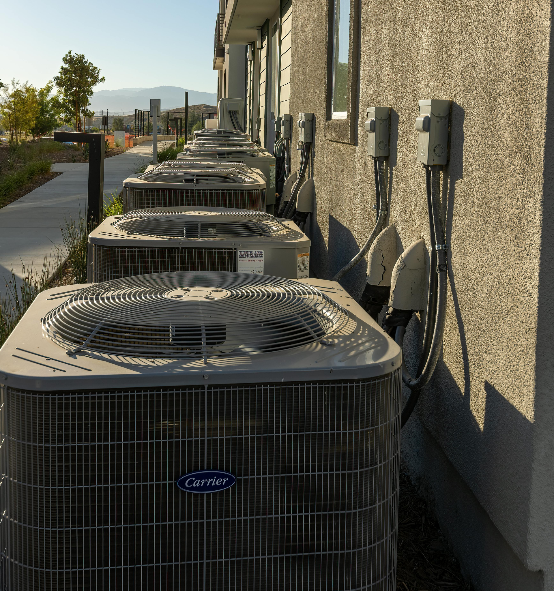 Row of outdoor air conditioning units beside a beige building in bright sunlight