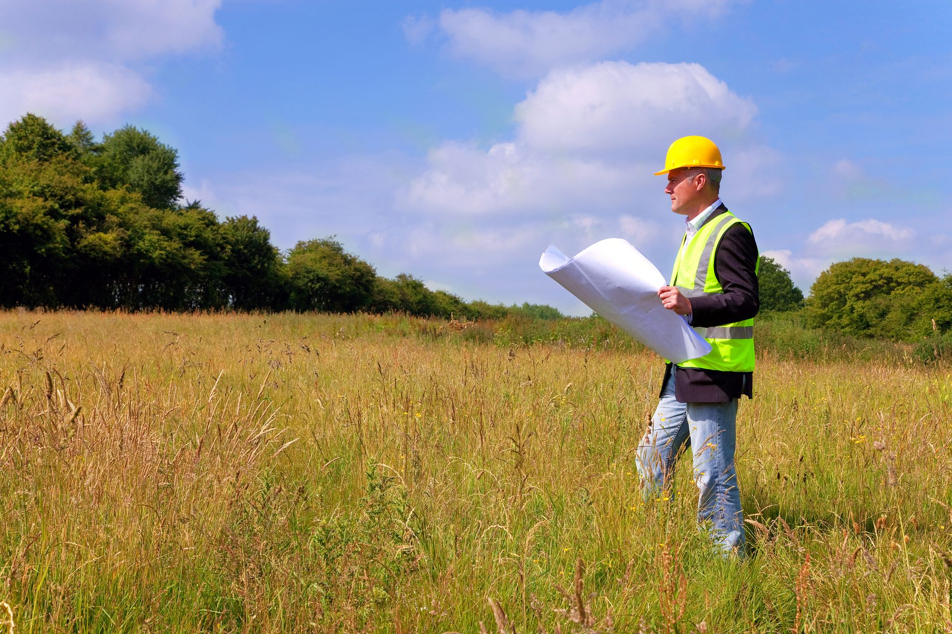 Surveyor in yellow hard hat and safety vest holding blueprints in a grassy field under a blue sky