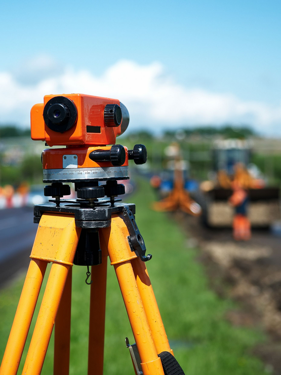 Orange surveying camera on yellow tripod outdoors, with blurred workers and field in the background