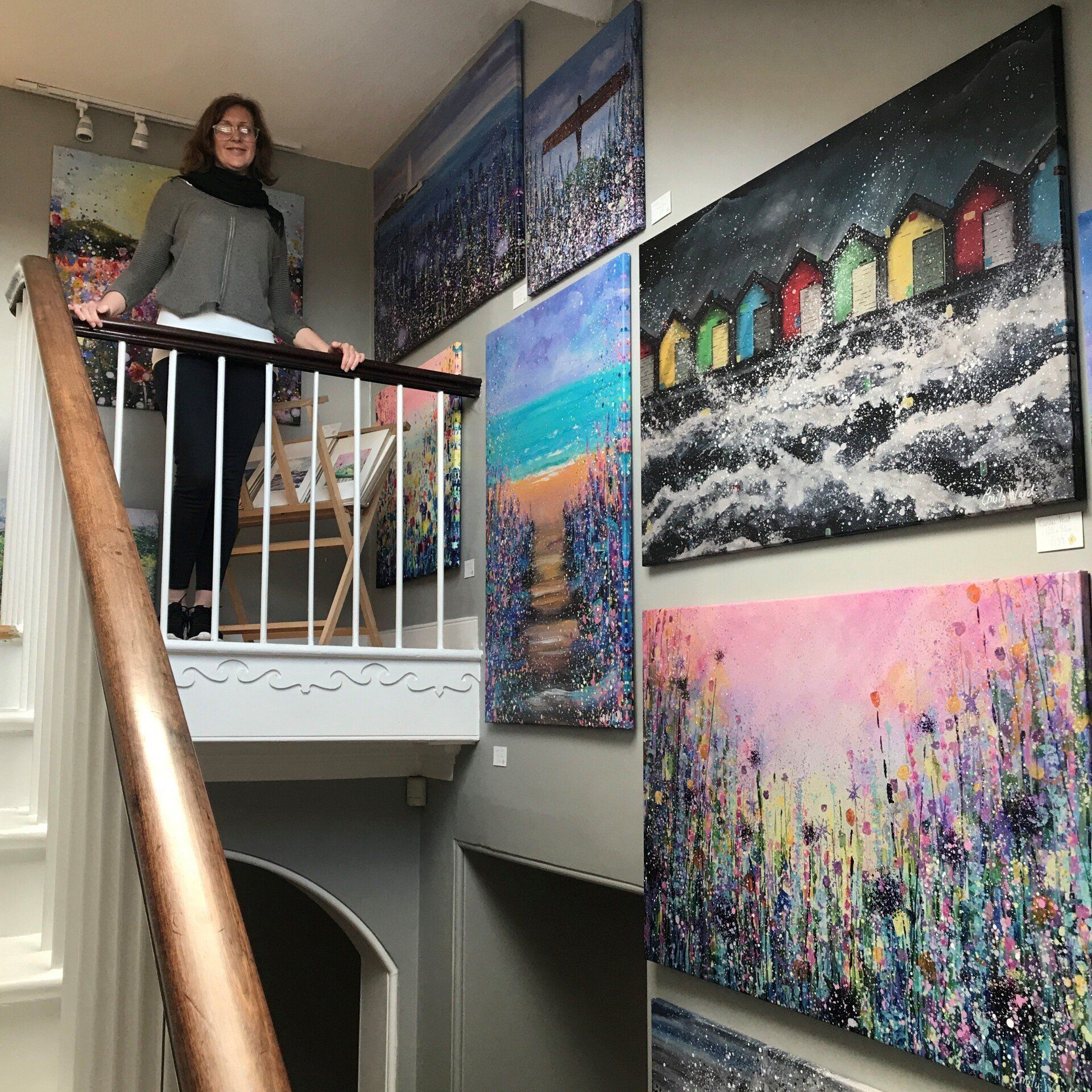 Woman standing on a stair landing beside colorful abstract paintings displayed on the walls.