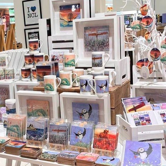 Colourful display of framed art prints, cards, and mugs arranged on a shop table.